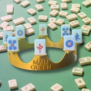 Peut inclure: Une couronne de couleur dorée portant l'inscription "MAHJ QUEEN" en relief sur le devant, avec plusieurs tuiles de mah-jong blanches fixées sur les pointes. Les tuiles présentent divers caractères chinois et motifs circulaires bleus et rouges.