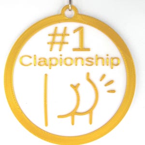May include: A gold-colored circular pendant with the words "#1 Clapionship" and a stylized graphic of a rear end. The pendant is suspended from a silver-colored metal loop.