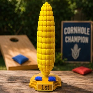 May include: A yellow corn-shaped trophy featuring a textured cob design mounted on a tiered base. The base displays the text 1st and includes two small blue bean bags resting on its surface, symbolizing a cornhole game victory award.