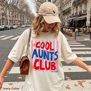 May include: An ivory t-shirt featuring a graphic print with vertical pink stripes and the text COOL AUNTS CLUB in red and blue, accented by orange stars and the text EST. 2026. The shirt is paired with a beige baseball cap and a brown shoulder bag.