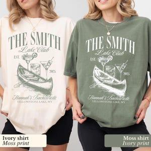 以下が含まれることがあります： カヌー、飲み物、そして「The Smith Lake Club Est. 2025」と「Hannah's Bachelorette Yellowstone Lake, WY」の文字が書かれたグラフィックデザインが施されたTシャツ2枚。左側のTシャツはアイボリーでモスグリーンのプリント。右側のTシャツはモスグリーンでアイボリーのプリントです。