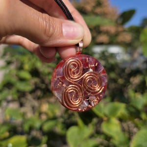 Könnte beinhalten: Ein runder Anhänger mit einer roten und violetten Steinbasis, verziert mit Kupferdrahtspiralen. Der Anhänger hängt an einem schwarzen Band. Das Design ist vor einem verschwommenen grünen und blauen Hintergrund platziert.
