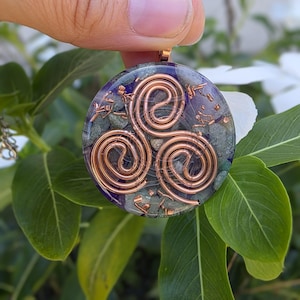 Könnte beinhalten: Runder Anhänger mit einem kupferfarbenen Spiraldesign auf einem Hintergrund aus blauen und grünen Steinen. Der Anhänger wird von einer kupferfarbenen Öse gehalten. Grüne Blätter und weiße Blüten umgeben den Anhänger.
