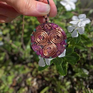 May include: A round pendant with a copper spiral design, set against a backdrop of crushed purple stones and green accents. The pendant is suspended from a black cord. The background features white flowers and green foliage.