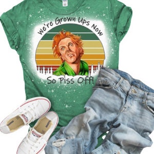 May include: Green t-shirt with a distressed, vintage-style graphic of a man with red hair and a green shirt. The graphic includes a sunset with a yellow, orange, and red color scheme. The text "We're Grown Ups Now So Piss Off!" is printed on the graphic.