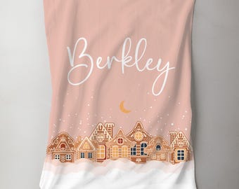 May include: A pink personalized blanket with the name "Berkley" in white cursive script. The blanket features a scene of gingerbread houses with white snow and a crescent moon.