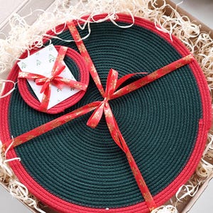 May include: A green and red braided placemat and coaster set, presented in a cardboard box with decorative raffia and a red ribbon. The set is a festive holiday gift, with a matching coaster and placemat.