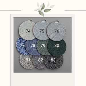 May include: A collection of nine round coasters in various colors, including gray, blue, and green. Each coaster has a loop for hanging and is numbered from 74 to 83. The coasters are arranged on a gray background.