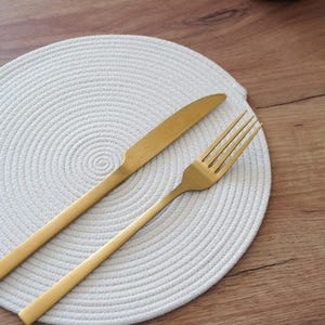 May include: A gold-colored knife and fork rest on a white, circular placemat with a woven texture. The flatware is positioned diagonally across the mat, set against a wooden table. A red and white mug is visible in the background.