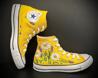 May include: A pair of bright yellow high-top sneakers with white laces and soles. The sneakers are adorned with embroidered daisies and lavender sprigs. The Converse logo is visible on the side. The shoes are approximately 6 inches tall.