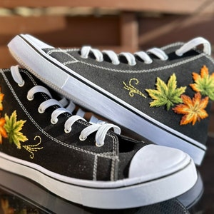 Op de afbeelding: Zwarte high-top sneakers met witte zolen en veters. De sneakers hebben geborduurde esdoornbladontwerpen in groen, geel en oranje. De schoenen hebben een klassiek canvas ontwerp.