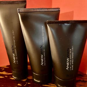 May include: Three black tubes of ANEW MEN skincare products. The tubes are labeled with white text, including "2-IN-1 GEL CLEANSER", "PROTECTIVE SHAVE GEL", and "2-IN-1 AFTER SHAVE BALM". The tubes have black caps and are arranged vertically.