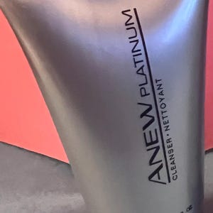 May include: A silver tube of ANEW PLATINUM cleanser, with the text "CLEANSER NETTOYANT" and "50 ml 1.7 FL OZ". The tube has a black cap and is set against a pink and red background.