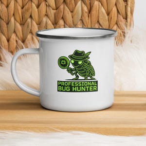 Op de afbeelding: Een witte emaille mok met een zilveren rand en een groene afbeelding van een insect met een hoed op die een vergrootglas vasthoudt. Onder de illustratie staat de tekst PROFESSIONAL BUG HUNTER in dikke groene hoofdletters op een donkere achtergrond.