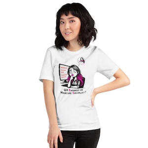 May include: A white short-sleeved t-shirt featuring a graphic illustration of a person with pink-streaked hair sitting at a computer. The text below reads "QA ENGINEER OR MANICURE SPECIALIST?" with the letters E and S highlighted in pink.