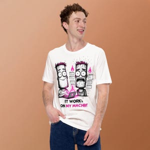 Peut inclure: Un t-shirt blanc à manches courtes avec une illustration graphique de deux personnages de dessin animé aux cheveux roses, l'un souriant devant un ordinateur portable et l'autre stressé. Le texte indique IT WORKS ON MY MACHINE.