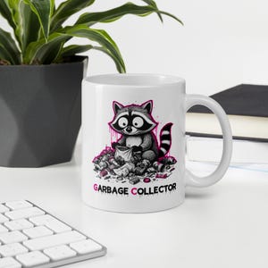 May include: A white ceramic mug featuring a graphic illustration of a raccoon sitting amidst a pile of trash. The raccoon is holding a small bag. Below the illustration, the text GARBAGE COLLECTOR is printed in black with pink accents.