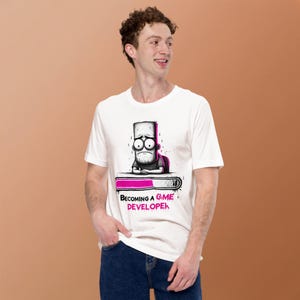 May include: A white short-sleeved t-shirt featuring a black and white cartoon illustration of a stressed character sitting at a desk above a pink loading bar. Below the graphic, the text reads "Becoming a GAME DEVELOPER" in black and pink.