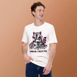 Peut inclure: Un t-shirt blanc à manches courtes orné d'une illustration graphique représentant un raton laveur assis dans un tas d'ordures. Le raton laveur tient un petit sac. Sous le dessin, le texte GARBAGE COLECTOR est écrit en lettres noires.