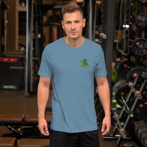 Op de afbeelding: Een lichtblauw t-shirt met korte mouwen en een kleine afbeelding op de linkerborst. De afbeelding toont een groen cartoon-insect met een hoed en een vergrootglas, met de tekst "PROFESSIONAL BUG HUNTER" eronder gedrukt.