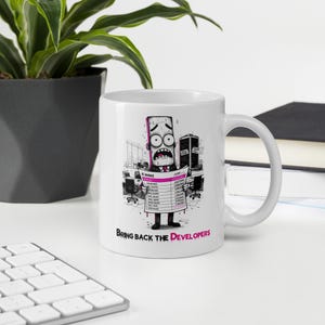 May include: A white ceramic mug featuring a cartoon illustration of a stressed, sweating character in a suit holding an AI invoice. The graphic includes office furniture and the text "BRING BACK THE DEVELOPERS" in black and pink lettering.
