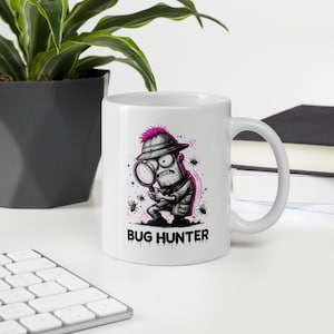 Può includere: Una tazza in ceramica bianca con un'illustrazione a fumetti in bianco e nero di un personaggio che indossa un casco coloniale e tiene una lente d'ingrandimento, circondato da piccoli insetti, con il testo BUG HUNTER in grassetto nero.