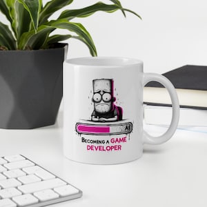 May include: A white ceramic mug featuring a black and white illustration of a stressed character leaning on a loading bar. The loading bar is partially filled with pink. Below the graphic, the text reads "BECOMING A GAME DEVELOPER" in black and pink.