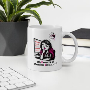 Pode incluir: Uma caneca de cerâmica branca com uma ilustração gráfica de uma pessoa com cabelo escuro e madeixas cor-de-rosa sentada ao computador. Acima da pessoa, um balão de pensamento mostra uma manicure. O texto diz: QA ENGINEER OR MANICURE SPECIALIST?