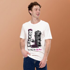 Puede incluir: Una camiseta blanca de manga corta con una ilustración gráfica de un personaje de dibujos animados con pelo rosa usando un joystick para controlar un rack de servidores envuelto en cables y bombas, con el texto "Let's Test It On Pro".