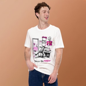 Può includere: Una t-shirt bianca a maniche corte con un'illustrazione grafica di un personaggio dai capelli rosa alla scrivania, che preme un pulsante con l'etichetta DEPLOY. La scena include una vista sulla spiaggia, un calendario segnato FRIDAY e il testo DEPLOY ON FRIDAY.