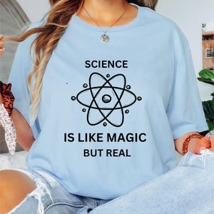 May include: Light blue t-shirt with the text "SCIENCE IS LIKE MAGIC BUT REAL" and a black atom graphic. The shirt is made of a soft material and is suitable for casual wear. The shirt is a crew neck style.