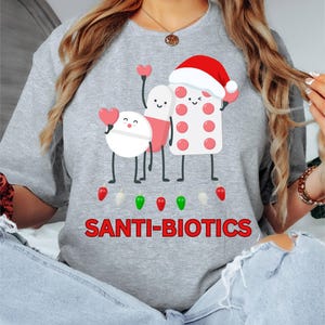 Puede incluir: Una sudadera gris con el texto "SANTI-BIOTICS" en rojo. La camiseta presenta ilustraciones de dibujos animados de pastillas y un blíster, algunos con corazones y gorros de Papá Noel. Debajo del texto hay ilustraciones de luces navideñas.