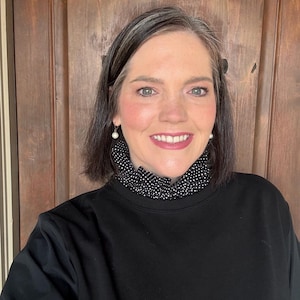 May include: A woman wearing a black sweatshirt with a black and white polka dot collar. She is wearing pearl earrings. The background is a wooden door.