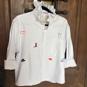 May include: A white long-sleeve button-down shirt with a ruffled collar. The shirt features embroidered details including a cowboy boot, a hat, the text "pistols firing", a bow, the state of Oklahoma, and the text "est 1890" in orange and black.
