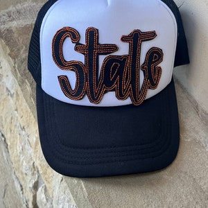 May include: A black and white trucker hat with a black mesh back. The front of the hat has a white panel with the word "State" in black with orange stitching.