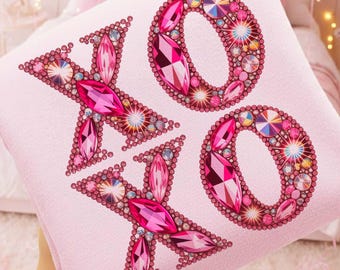 May include: A light pink throw blanket with the letters "XO" embellished with pink and iridescent rhinestones. The blanket is held up in front of a bed with pink balloons in the background. The blanket is a decorative item.