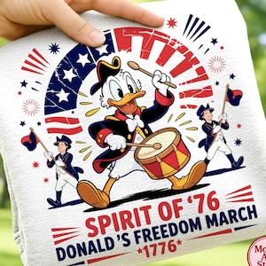 Könnte beinhalten: Weißes dekoratives Handtuch mit Donald Duck, der in patriotischer Kleidung Trommel spielt, mit dem Text "Spirit of '76 Donald's Freedom March *1776*". Das Design umfasst rote, weiße und blaue Elemente, Sterne und Illustrationen von marschierenden Figuren.