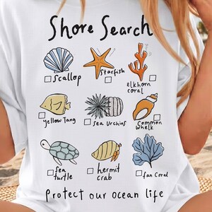 May include: White t-shirt with the words "Shore Search" and illustrations of sea creatures like a scallop, starfish, and sea turtle. The text "Protect our ocean life" is also printed on the shirt. A bottle of Coca-Cola is held in the left hand.