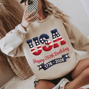 May include: Beige sweatshirt with the word "USA" in red, white, and blue, celebrating the 250th birthday of the United States. The sweatshirt also features the years 1776-2026 on a banner.