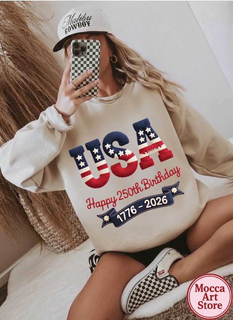 May include: Beige sweatshirt with the word "USA" in red, white, and blue, celebrating the 250th birthday of the United States. The sweatshirt also features the years 1776-2026 on a banner.