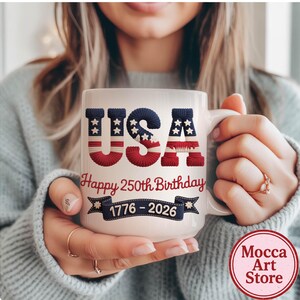 May include: White ceramic mug featuring the word "USA" in red, white, and blue, with a stars and stripes design. The mug also has the text "Happy 250th Birthday" and "1776-2026". The mug is held by a person.