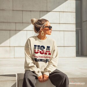 May include: A cream-coloured sweatshirt featuring the word "USA" in blue and red, commemorating the 250th birthday from 1776-2026. The casual sweatshirt is suitable for patriotic events or everyday wear.