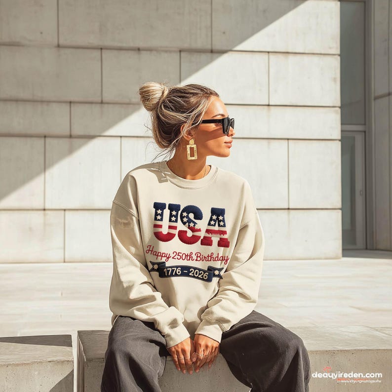 May include: A cream-coloured sweatshirt featuring the word "USA" in blue and red, commemorating the 250th birthday from 1776-2026. The casual sweatshirt is suitable for patriotic events or everyday wear.