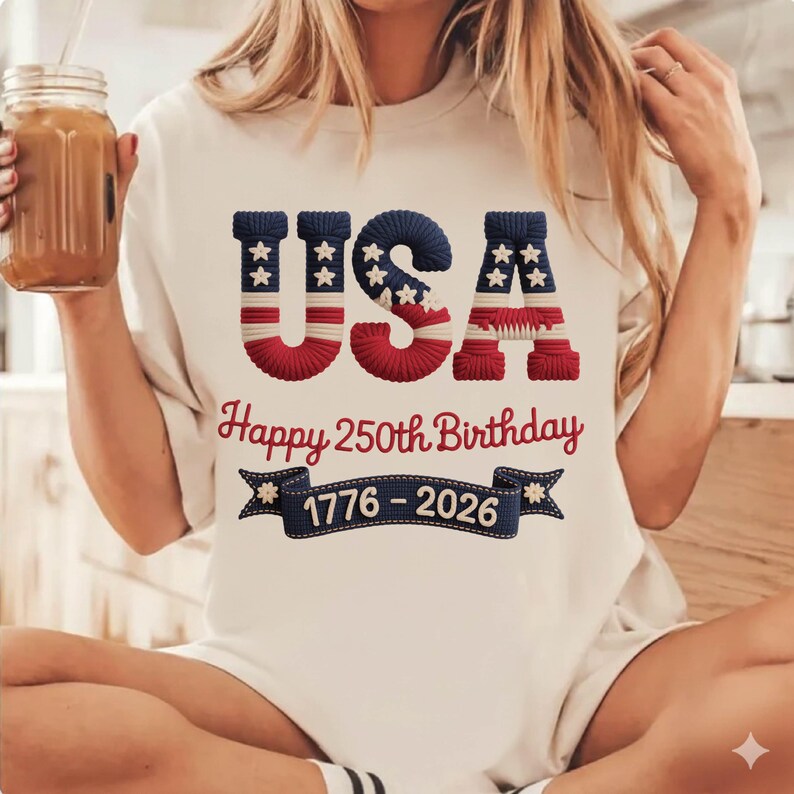 May include: Cream-colored t-shirt with the word "USA" in blue and red, with white stars. Below, the text reads "Happy 250th Birthday" and "1776-2026" on a banner. The shirt is a patriotic design.
