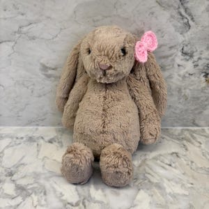 Peut inclure: Un lapin en peluche de couleur beige avec de longues oreilles tombantes et un nœud rose. La peluche a un aspect doux et texturé et est assise. Le nez du lapin est rose clair.