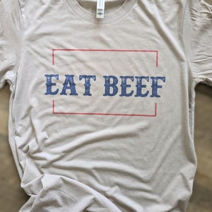 May include: A light gray t-shirt with a red and white border around the text "EAT BEEF" in blue.