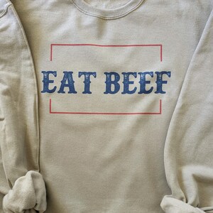 May include: A light beige sweatshirt with the text "EAT BEEF" printed in blue letters inside a red rectangle.