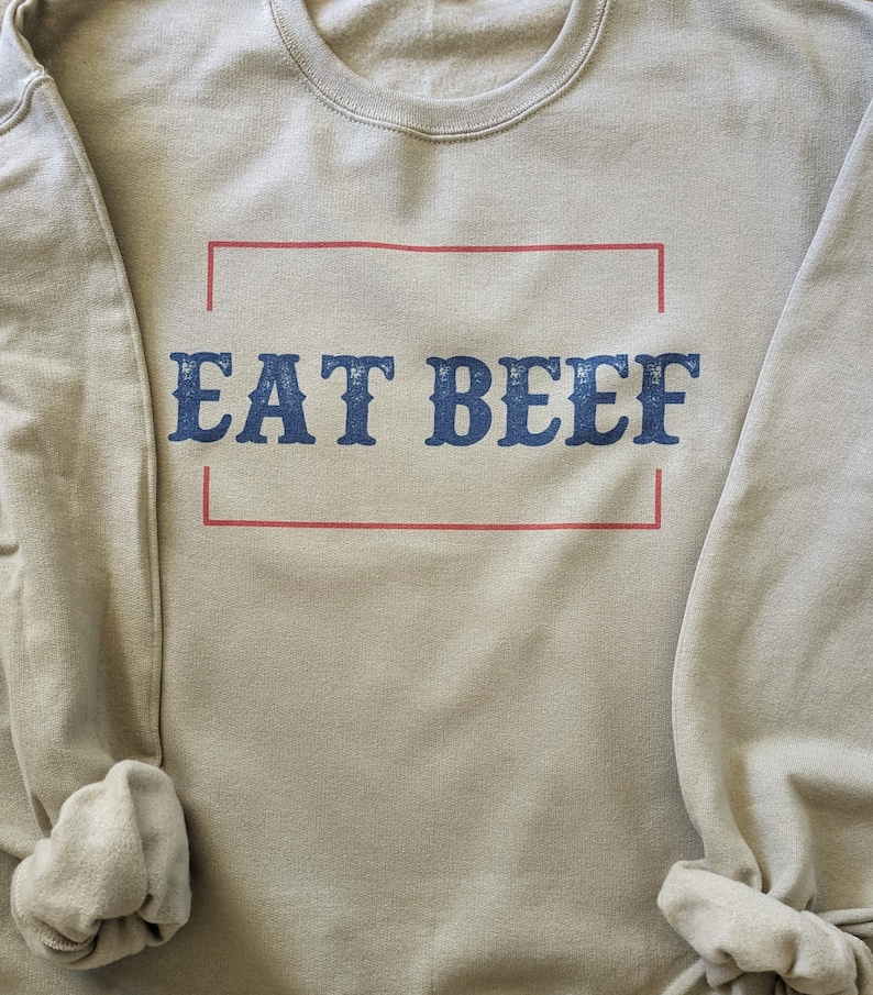 May include: A light beige sweatshirt with the text "EAT BEEF" printed in blue letters inside a red rectangle.