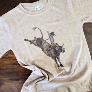 May include: A tan t-shirt with a graphic of a cowboy riding a bucking bull. The bull is brown and black and the cowboy is wearing a brown hat and a light blue shirt.