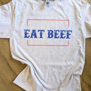 May include: A gray t-shirt with the text "EAT BEEF" printed in blue letters inside a red rectangle.